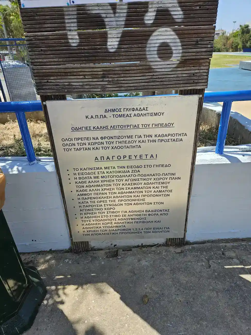 Ξύλινη κατασκευή με ανακοίνωση του Δήμου Γλυφάδας για τους κανόνες και τις ώρες λειτουργίας του στίβου, με φόντο μπλε κάγκελα