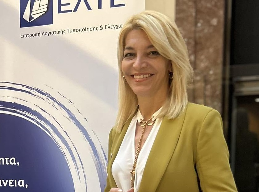 Η Αφροδίτη Νέστορα με λαδί κοστούμι δίπλα στο banner της Επιτροπής Λογιστικής Τυποποίησης και Ελέγχων